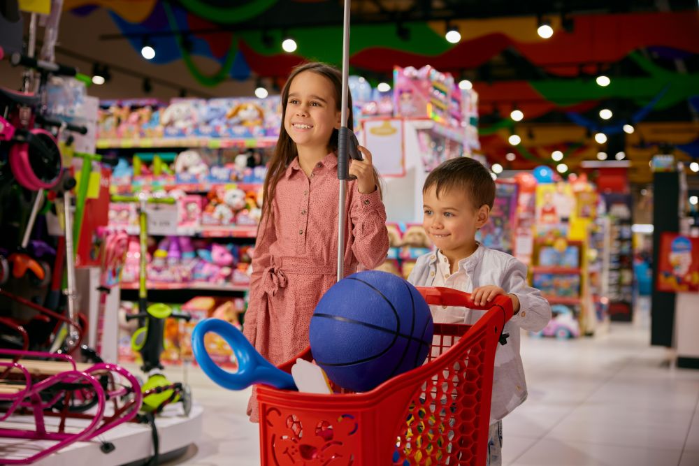 Mädchen und Bube fahren mit einem kleinen Einkaufswagen durch einen Spielzeughandel